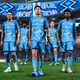 Some foot-to-ball players in blue, walking on the pitch.